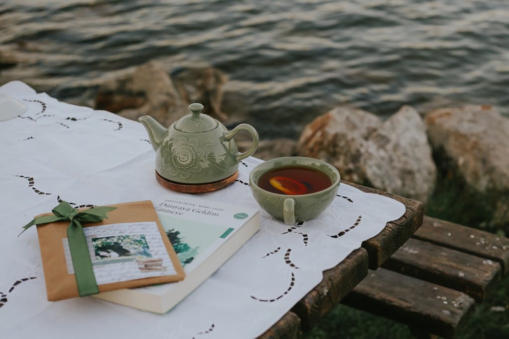A serene lakeside table with a teapot, cup of tea, book, and wrapped gift on a lace tablecloth.